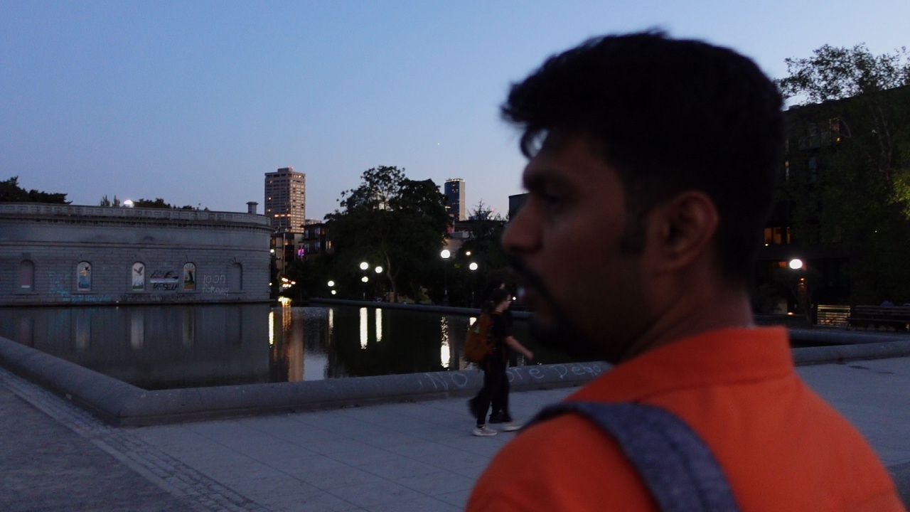 SUSH 28 in profile at dusk beside a reflecting pool, with city buildings behind him